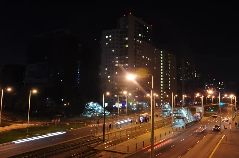 Avenida iluminada à noite, com rastros de luz dos carros e prédios residenciais ao fundo