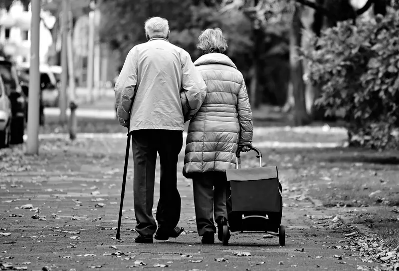 Casal de idosos caminhando de costas por uma calçada arborizada, em preto e branco