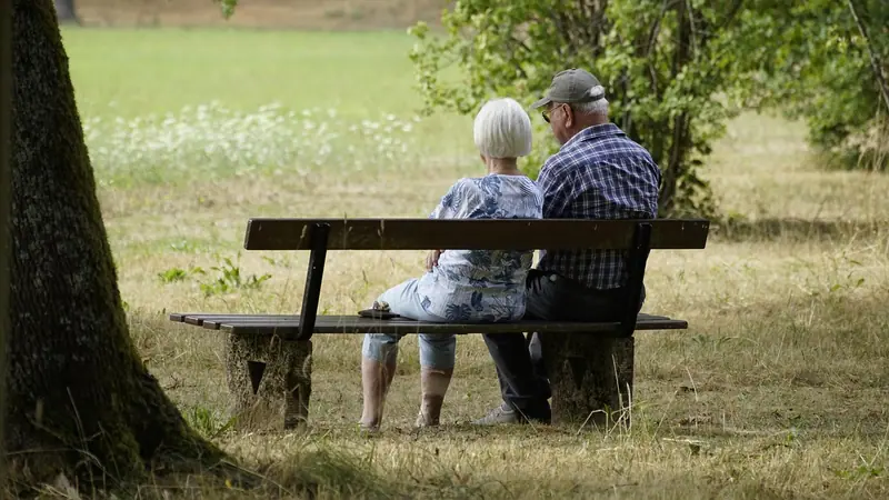 Casal de idosos sentado de costas num banco de praça, à sombra de uma árvore