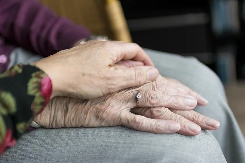 Mão de uma pessoa mais jovem pousada sobre as mãos de uma idosa, apoiadas no colo