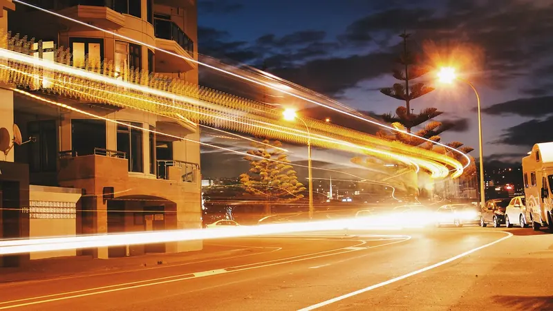 Rastros de luz dos carros numa avenida à noite, passando rente a um prédio residencial iluminado