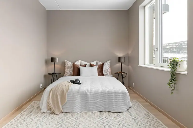 Quarto claro com cama de casal arrumada, almofadas, luminárias e uma janela ampla com vista para fora