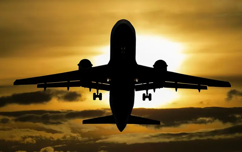 Silhueta de um avião de passageiros subindo diante do sol, com o céu alaranjado