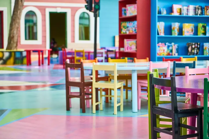 Mesas e cadeirinhas coloridas num espaço infantil vazio, com estantes de brinquedos ao fundo