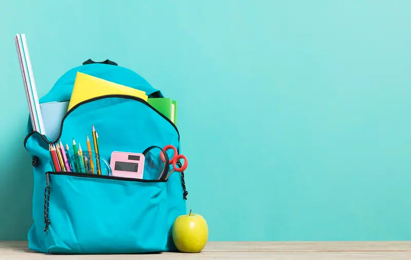 Mochila azul cheia de material escolar, com lápis, régua e tesoura, ao lado de uma maçã verde
