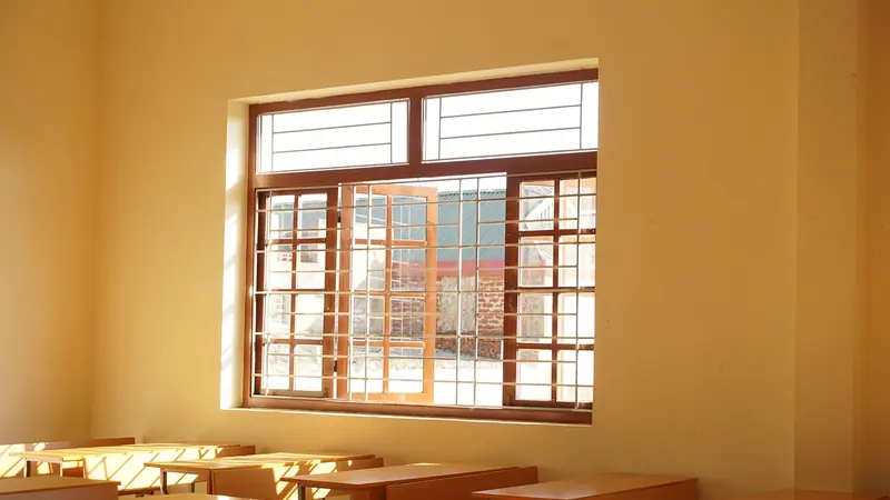 Sala de aula vazia com carteiras de madeira e uma janela grande gradeada iluminada pelo sol