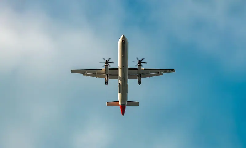Avião visto de baixo, com as rodas abertas, cruzando um céu azul com nuvens