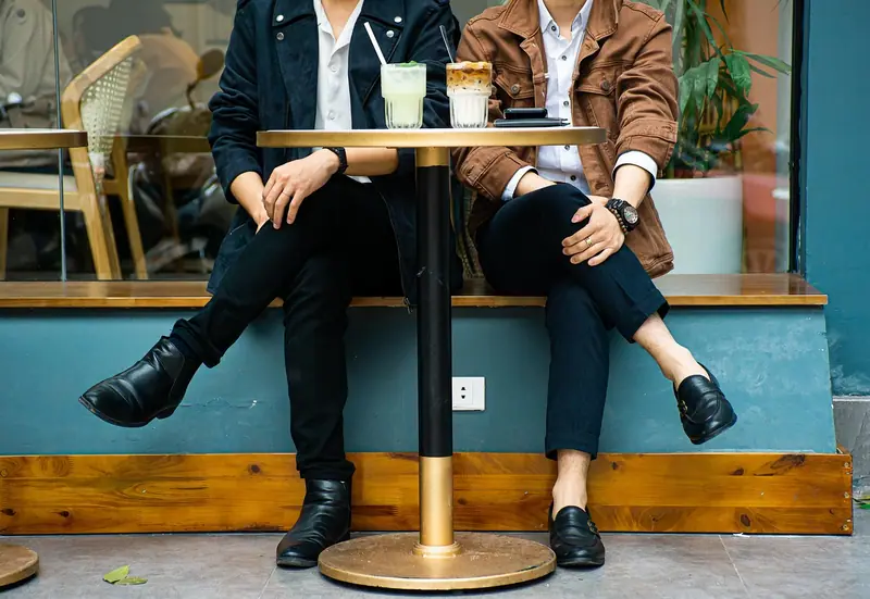 Duas pessoas sentadas a uma mesa alta de bar, vistas só dos joelhos para baixo, com copos sobre a mesa