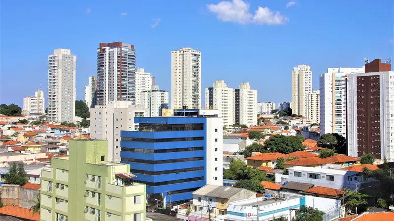 Bairro de São Paulo com prédios altos atrás de casas de telhado vermelho, sob céu azul com nuvens