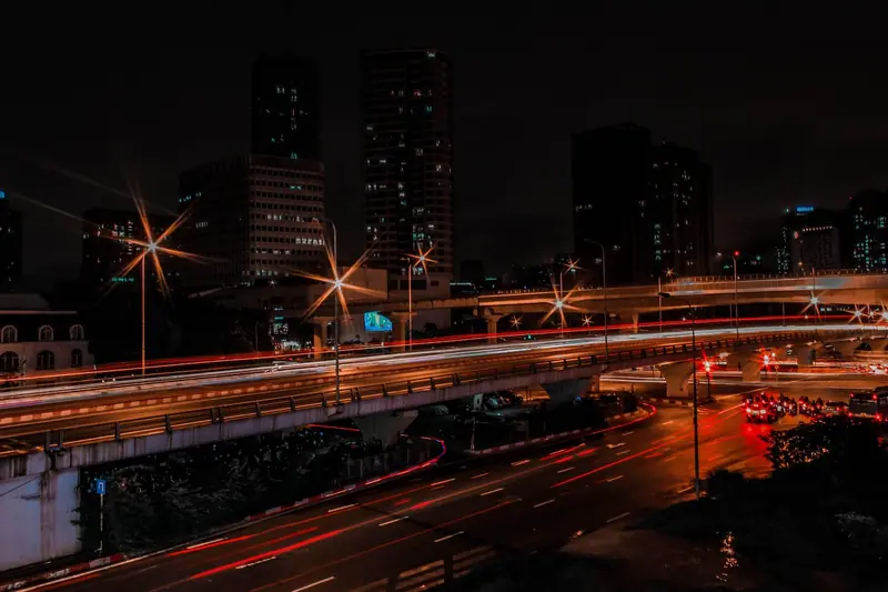 Viaduto à noite com rastros de luz dos carros e prédios iluminados ao fundo