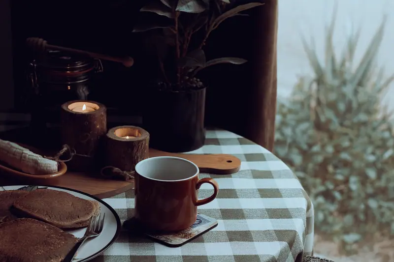 Xícara, velas acesas e uma planta sobre uma mesa com toalha xadrez, à meia-luz