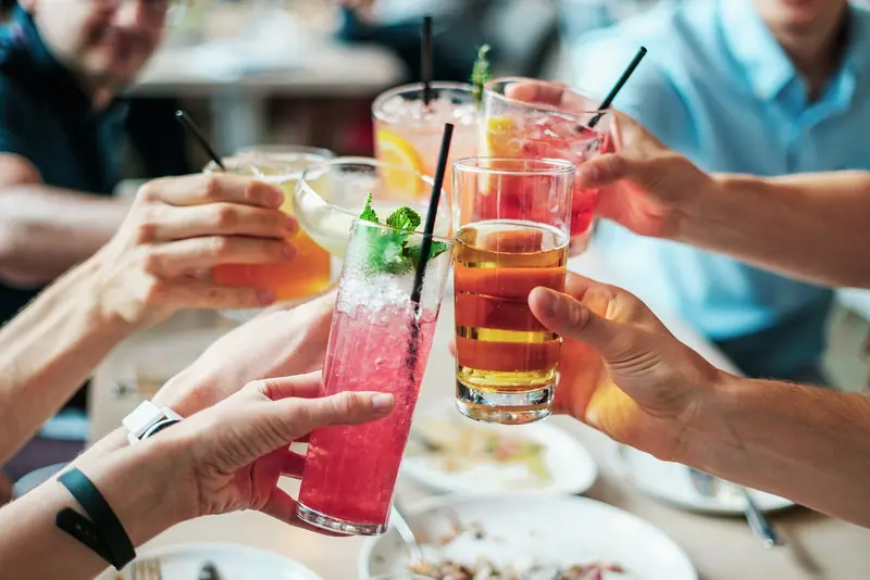 Mãos de várias pessoas brindando com copos de drinques coloridos numa mesa