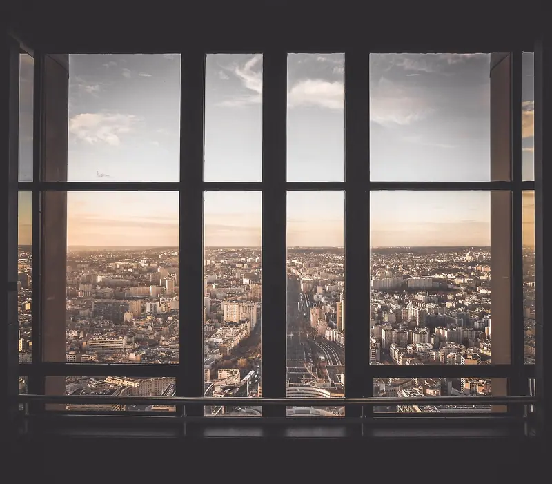 Janela ampla de um andar alto com vista para uma cidade cheia de prédios ao entardecer