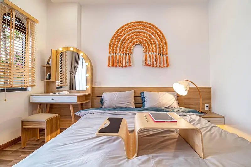 Quarto claro com cama arrumada, penteadeira e luminária ao lado da janela