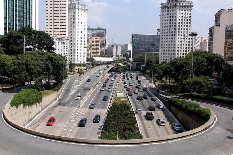 Avenida larga com carros nos dois sentidos, fazendo uma curva entre prédios altos e árvores