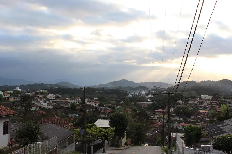 Rua de bairro com casas, árvores e fios de energia ao entardecer, com morros ao fundo