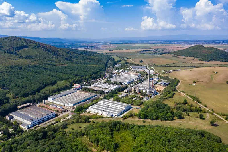 Galpões industriais de telhado claro vistos do alto, ao lado de morros com mata e campos