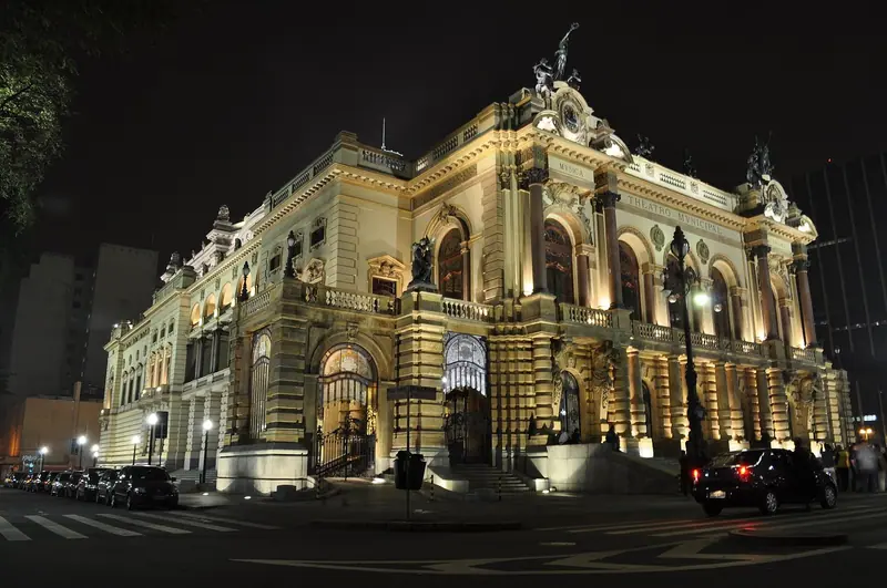 Teatro histórico iluminado à noite no centro de São Paulo, com carros passando na rua em frente