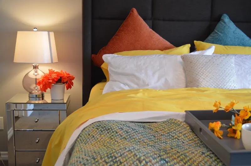 Cama com almofadas coloridas, manta amarela e abajur aceso sobre a mesa de cabeceira