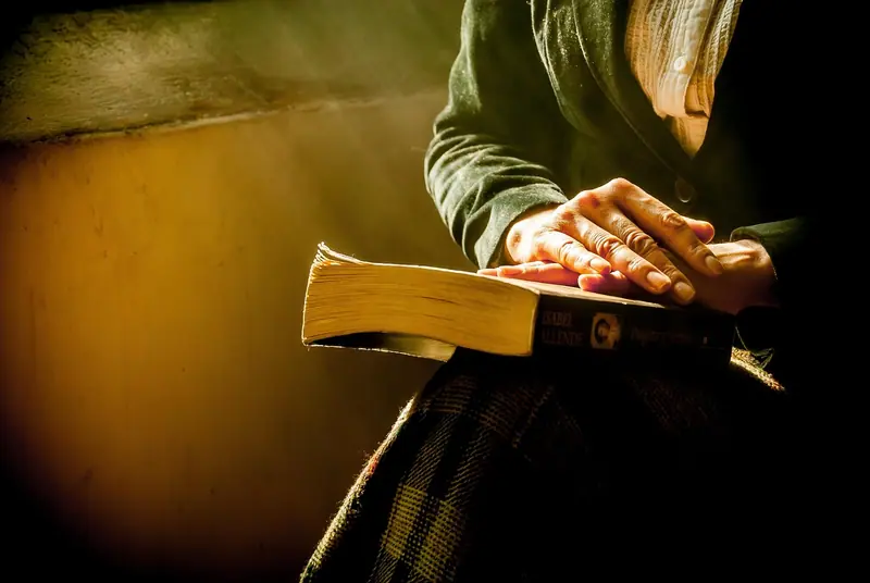 Mãos apoiadas sobre um livro aberto no colo, num ambiente de luz suave