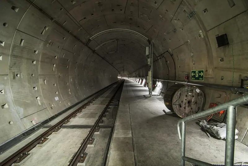 Túnel de concreto em construção, com trilhos provisórios, iluminação e equipamentos ao fundo