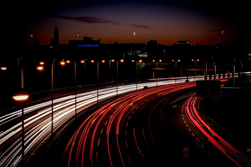 Via expressa em curva à noite, com rastros de luz dos faróis e postes acesos