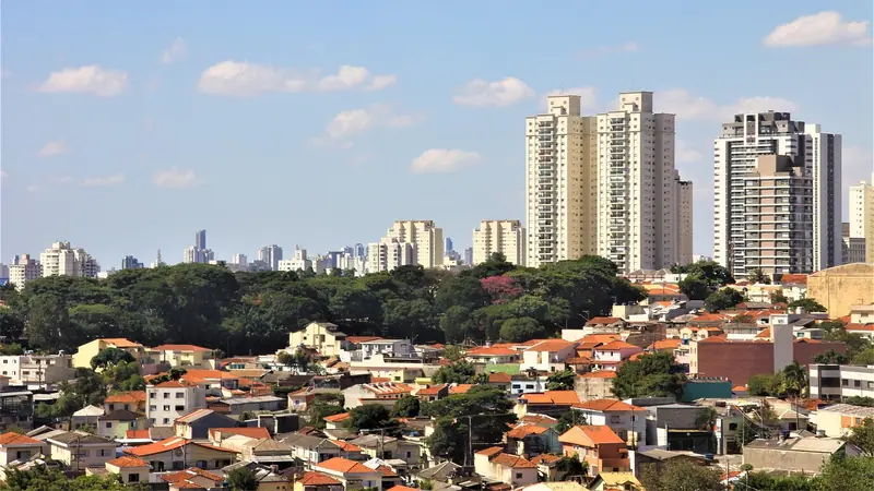 Casas de telhado vermelho e prédios altos ao fundo, entre árvores, sob céu azul com nuvens