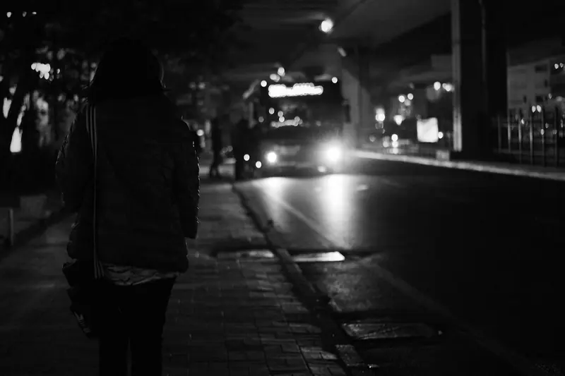 Pessoa de costas num ponto de ônibus à noite, com um ônibus de faróis acesos chegando, em preto e branco