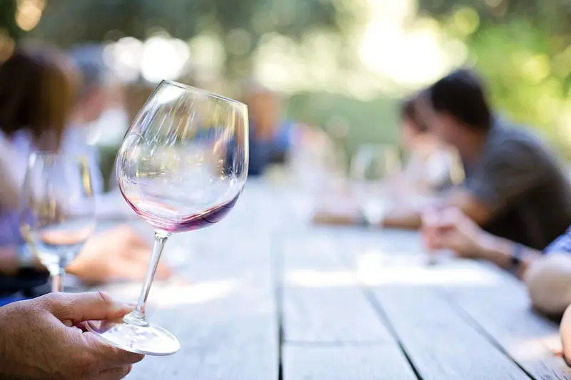 Mão segurando uma taça de vinho numa mesa de madeira ao ar livre, com pessoas conversando ao fundo