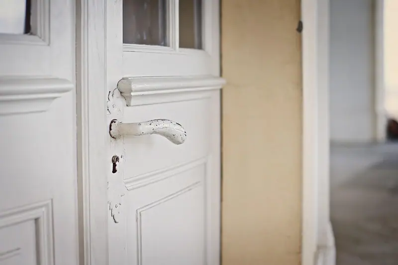 Porta branca antiga com a pintura gasta e maçaneta descascada, entreaberta para um corredor