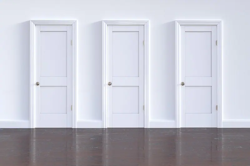 Três portas brancas fechadas lado a lado numa parede branca com piso de madeira