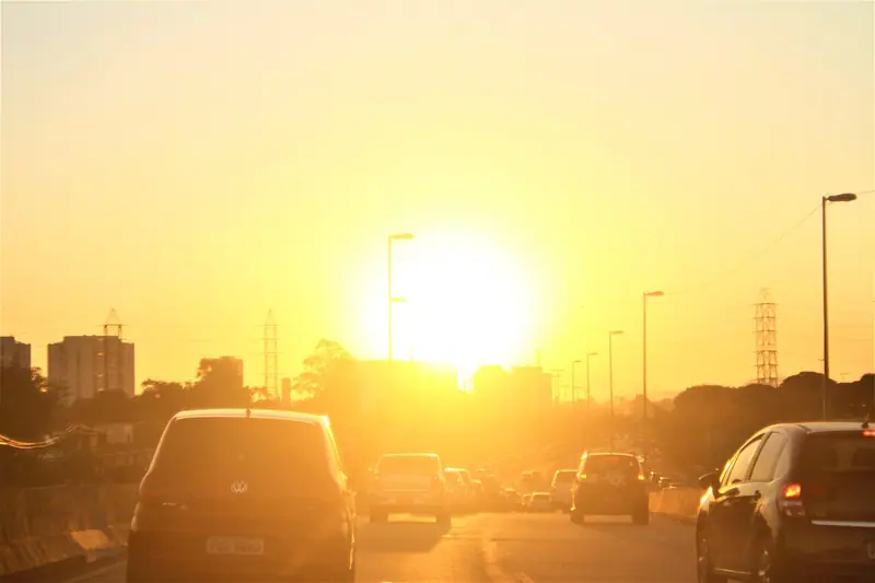 Avenida com carros parados no trânsito ao entardecer, com o sol baixo entre postes e prédios