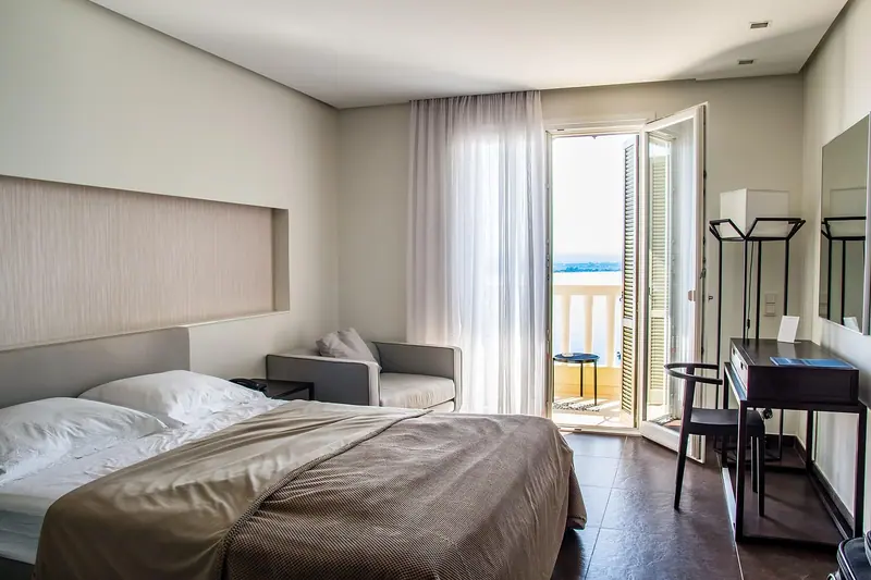 Quarto com cama de casal, escrivaninha e porta de varanda aberta com cortinas leves
