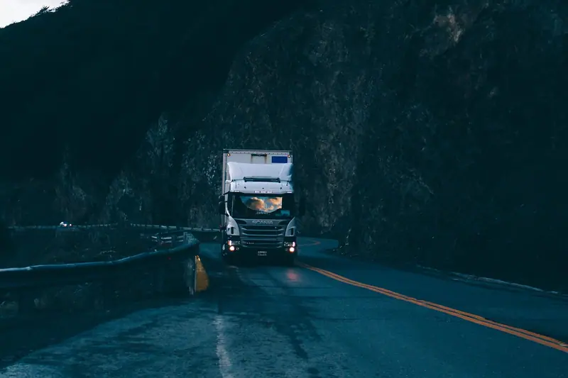 Caminhão baú com os faróis acesos numa estrada escura, ao lado de um paredão de pedra