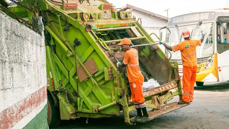 Dois garis de uniforme laranja na traseira de um caminhão de lixo verde, com um ônibus parado atrás