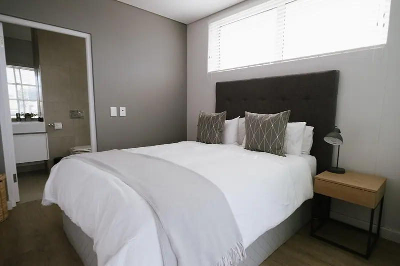 Quarto claro com cama de casal arrumada, cabeceira escura e uma janela alta com persiana