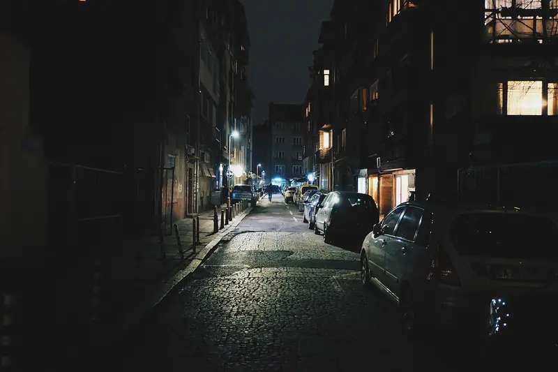 Rua estreita e escura de madrugada, com carros estacionados e poucas janelas acesas nos prédios