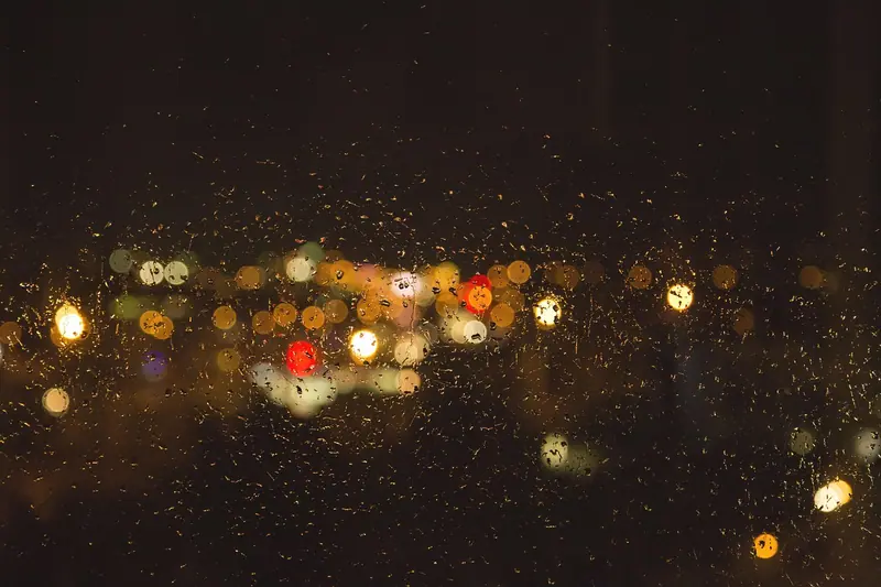 Luzes da cidade desfocadas atrás de um vidro com gotas de chuva, à noite