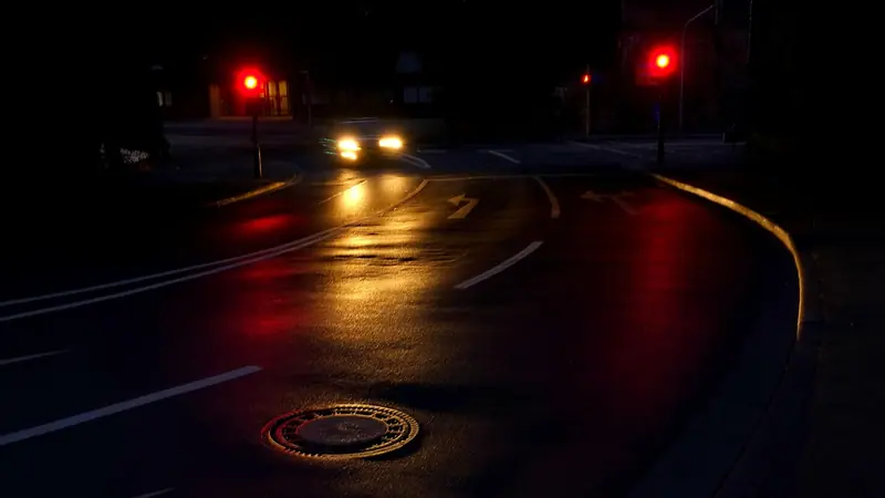 Rua escura e molhada à noite, com um carro de faróis acesos ao fundo e semáforos vermelhos