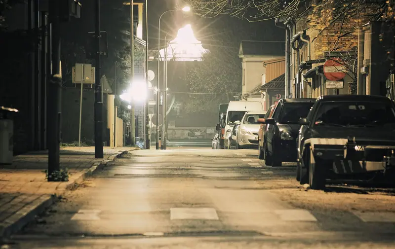 Rua residencial à noite, iluminada por postes, com carros estacionados junto à calçada