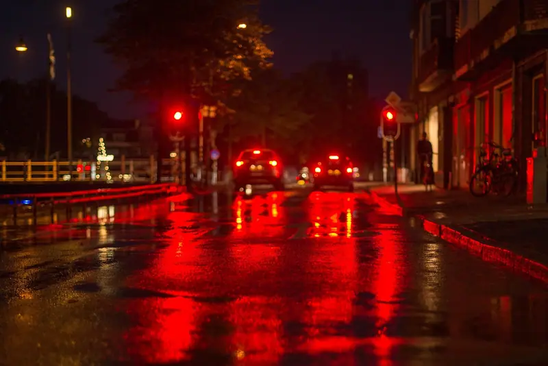 Carros parados no semáforo vermelho numa rua molhada à noite, com o asfalto refletindo as luzes