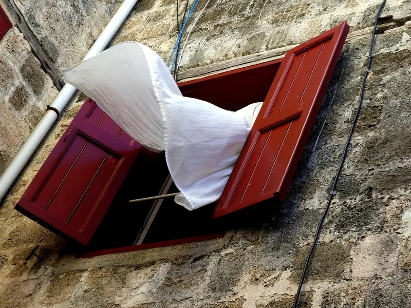 Cortina branca saindo ao vento por uma janela aberta de venezianas vermelhas, numa parede de pedra