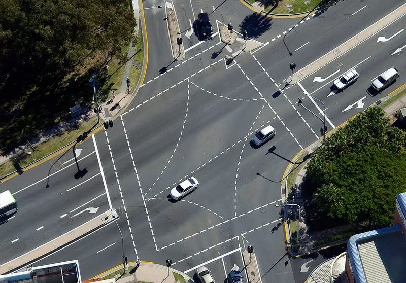 Cruzamento de duas avenidas visto do alto, com faixas pintadas, setas de conversão e carros fazendo a curva