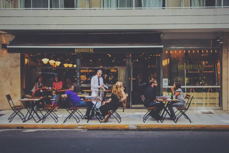 Mesas e cadeiras de um bar na calçada, com pessoas sentadas conversando em frente à fachada envidraçada