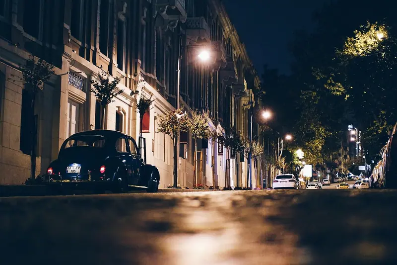 Rua de prédios antigos à noite, iluminada por postes, com um fusca preto estacionado junto à calçada