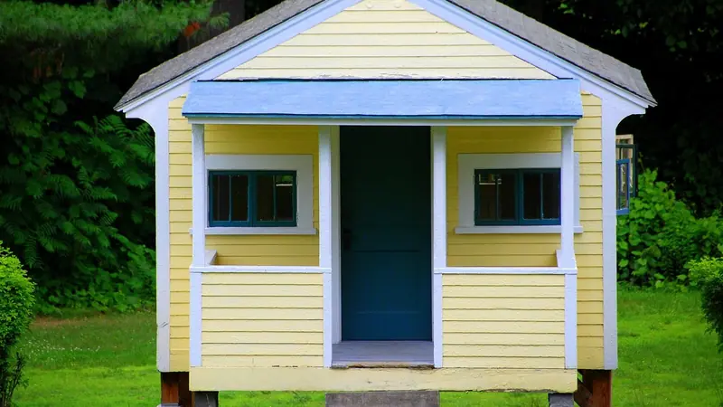Casinha amarela de madeira com porta azul e duas janelas pequenas, num gramado cercado de árvores
