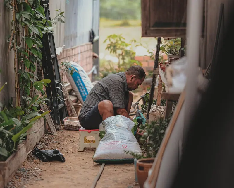Homem agachado num corredor lateral estreito de terra, entre a parede da casa e prateleiras com vasos