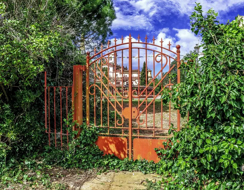 Portão de ferro trabalhado, pintado de laranja, fechado entre plantas e árvores