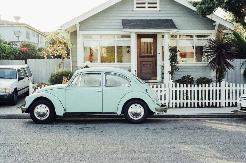 Fusca verde-claro estacionado na rua em frente a uma casa de varanda com cerca branca baixa