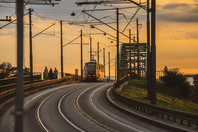 Bonde fazendo uma curva sobre trilhos numa ponte, ao pôr do sol, com fios elétricos sobre a via